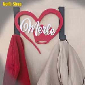 Könnte beinhalten: Ein rotes herzförmiges Holzschild mit dem Namen "Merle" in weißer Kursivschrift. Eine Krone befindet sich über dem Namen. Das Schild ist an einem schwarzen Metallhaken befestigt. Zwei Jacken hängen an den Haken.
