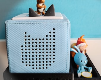 Puede incluir: Un altavoz azul claro con una rejilla de altavoz negra en forma de gato. El altavoz está sentado en un estante negro con el texto "Tonicbox" impreso en la parte delantera. Hay pequeñas figuras de plástico en la parte superior del altavoz.