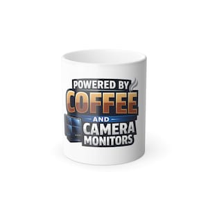 May include: A white ceramic mug featuring a graphic design with the text "POWERED BY COFFEE AND CAMERA MONITORS" in bold, multi-colored lettering. The design includes a small illustration of a multi-screen security camera display monitor.