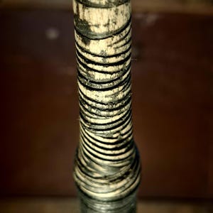 Op de afbeelding: Een cilindervormig handvat met een basis van lichtbeige hout, strak omwikkeld met een dun, donkerzwart koord. Het koord vormt een dicht, horizontaal spiraalpatroon dat het oppervlak van het handvat bedekt en zorgt voor een textuurrijke grip.