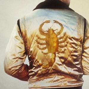 Puede incluir: Chaqueta bomber de satén dorado con cuello y ribetes negros. La parte trasera de la chaqueta presenta un gran emblema de escorpión dorado. La chaqueta tiene un patrón acolchado y un dobladillo acanalado.