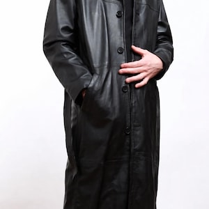 Op de afbeelding: Een lange, zwarte leren jas met een knoopsluiting en een kraag. De jas heeft zakken en een klassiek, getailleerd ontwerp. Het kledingstuk is gemaakt van een glad, glanzend materiaal en lijkt in uitstekende staat te verkeren.