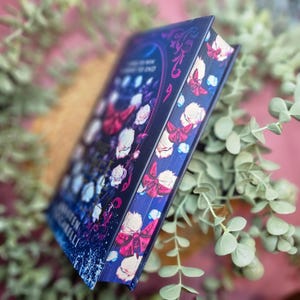 May include: A hardback book with a dark blue cover featuring a floral design with white roses, red butterflies, and gold accents. The title of the book is "The Cruel Prince" by Holly Black.