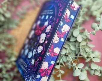 May include: A hardcover book with a dark blue cover featuring a floral design with white roses, red butterflies, and gold accents. The title of the book is "The Cruel Prince" by Holly Black.