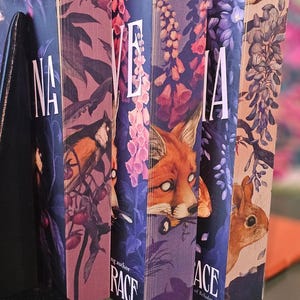 May include: A row of hardcover books with colorful covers. The spines display the title "NA", "VE", "IA", and "ACE" in white lettering. The covers feature illustrations of a bird, fox, and flowers. The book on the left has the title "Deliciously Deadly" by Stephanie Garber.