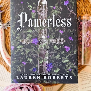 May include: A book cover with a dark green and black background featuring a silver sword with a purple flower on the hilt. The title "Powerless" is written in large, white letters. The author's name, "Lauren Roberts", is written in white letters below the title. The text "The Million Copy Bestselling Author" is written in white letters above the author's name.