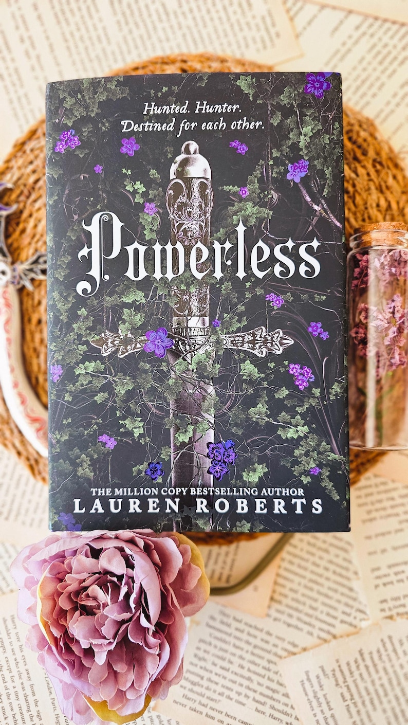 May include: A book cover with a dark green and black background featuring a silver sword with a purple flower on the hilt. The title "Powerless" is written in large, white letters. The author's name, "Lauren Roberts", is written in white letters below the title. The text "The Million Copy Bestselling Author" is written in white letters above the author's name.