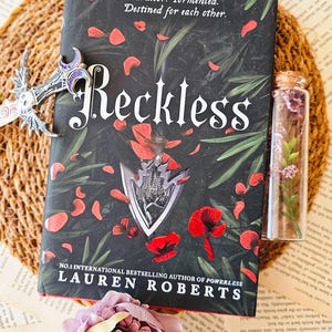 Può includere: Un libro tascabile intitolato "Reckless" di Lauren Roberts, l'autrice numero 1 di best seller internazionali di Powerless. La copertina presenta uno sfondo scuro con fiori rossi e foglie verdi. Il titolo è in testo bianco con un contorno nero. Il libro è appoggiato su una superficie intrecciata con una piccola bottiglia di vetro piena di fiori secchi e una chiave d'argento.