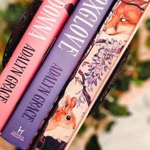 May include: Three hardcover books with titles "Belladonna" and "Foxglove" by Adalyn Grace. The books are in a decorative basket. The book covers are pink and purple with white lettering. The third book has a fox and rabbit illustration.