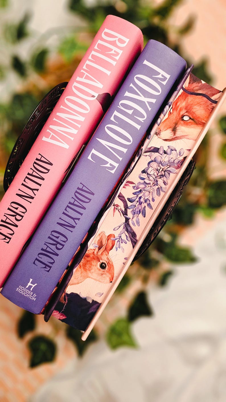 May include: Three hardcover books with titles "Belladonna" and "Foxglove" by Adalyn Grace. The books are in a decorative basket. The book covers are pink and purple with white lettering. The third book has a fox and rabbit illustration.