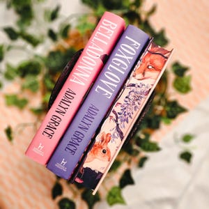 May include: Three hardcover books with the titles "Belladonna" and "Foxglove" are displayed. The books are in a dark brown basket. The book covers are pink and purple, with white text. The third book has a fox and floral design.