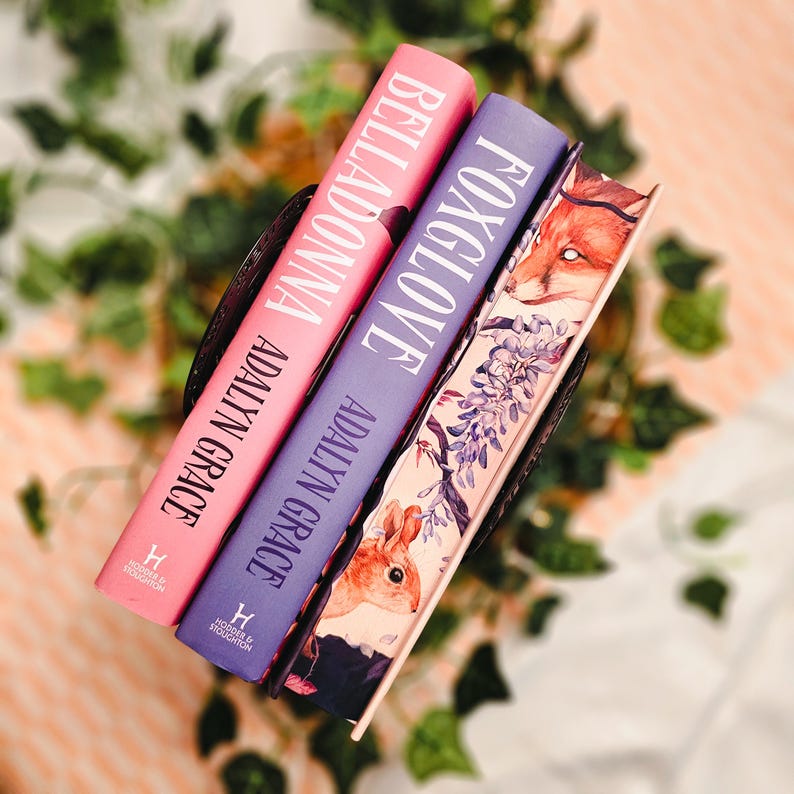 May include: Three hardcover books with the titles "Belladonna" and "Foxglove" are displayed. The books are in a dark brown basket. The book covers are pink and purple, with white text. The third book has a fox and floral design.