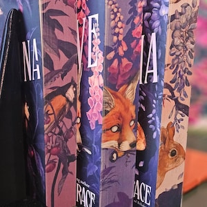May include: A row of hardcover books with colorful covers. The spines display the title "NA", "VE", "IA", and "ACE" in white lettering. The covers feature illustrations of a bird, fox, and flowers. The book on the left has the title "Deliciously Deadly" by Stephanie Garber.