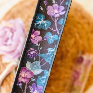 May include: Close-up of the spine of a hardcover book with a floral design. The design features purple and blue flowers on a black background.