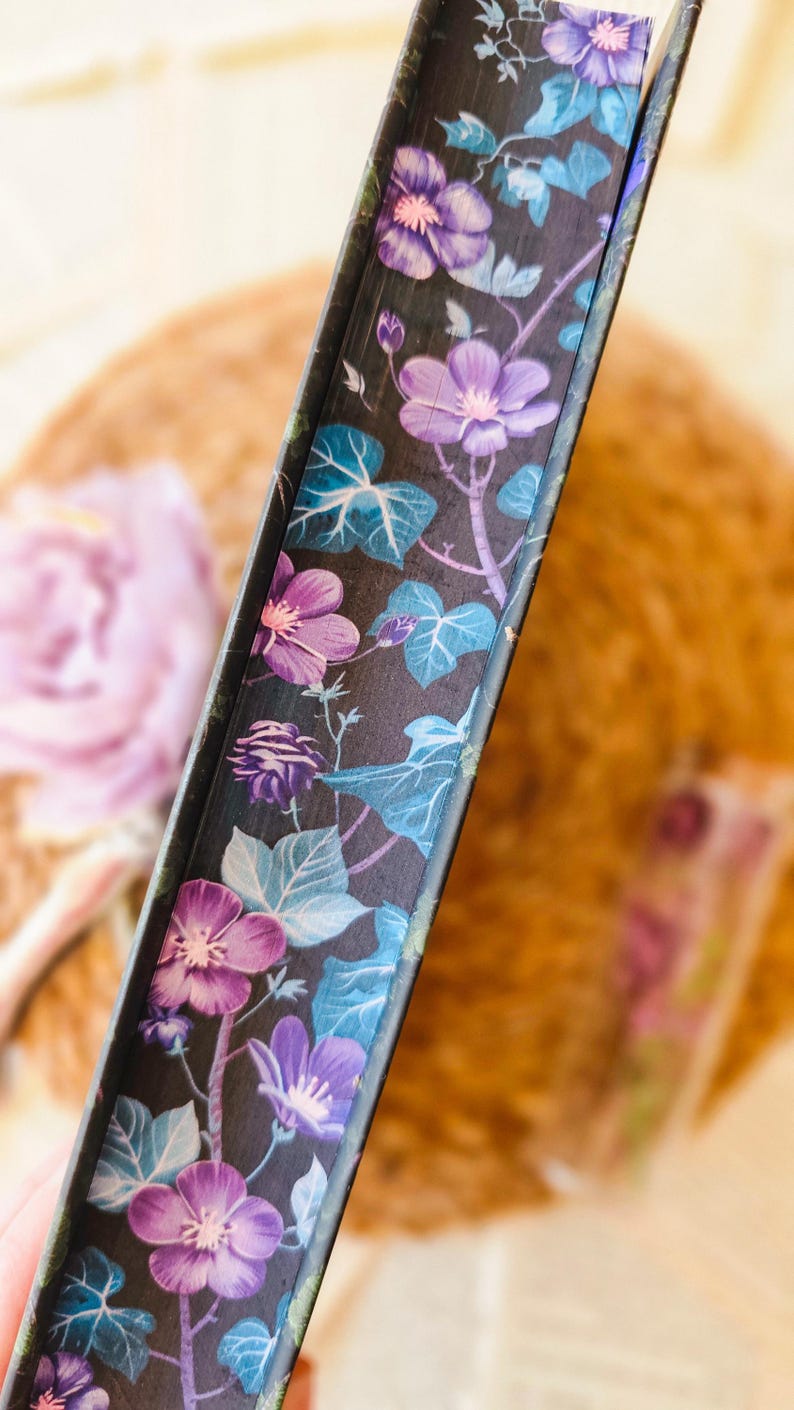 May include: Close-up of the spine of a hardcover book with a floral design. The design features purple and blue flowers on a black background.