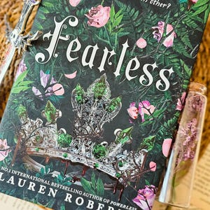 Könnte beinhalten: Ein Taschenbuch mit dem Titel "Fearless" von Lauren Roberts, der Nummer 1 internationalen Bestseller-Autorin von Powerless. Das Cover zeigt eine silberne Krone mit grünen Juwelen, umgeben von rosa und weißen Blumen und grünen Blättern auf schwarzem Hintergrund. Der Text "Betrothed. Betrayed. Destined for each other?" ist oben auf dem Cover in Weiß gedruckt.