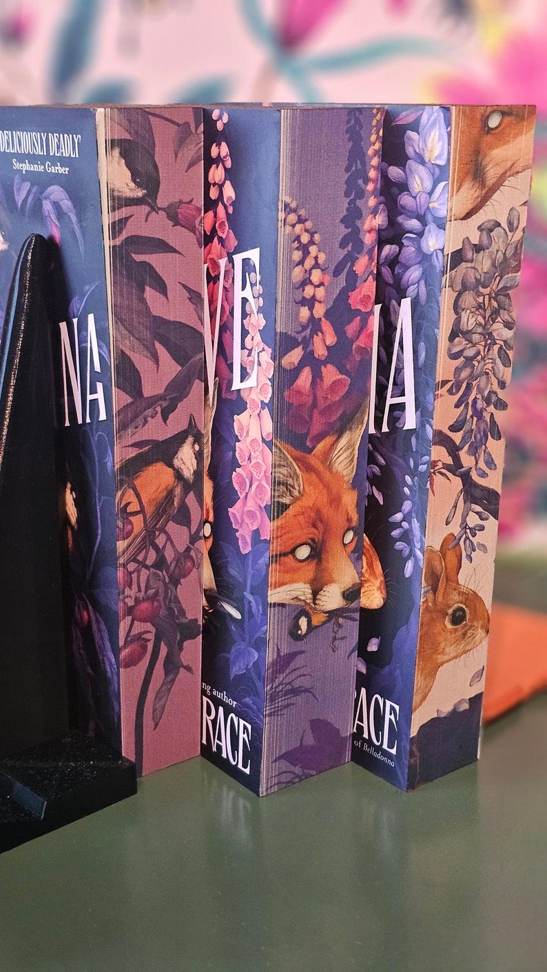 May include: A row of hardcover books with colorful covers. The spines display the title "NA", "VE", "IA", and "ACE" in white lettering. The covers feature illustrations of a bird, fox, and flowers. The book on the left has the title "Deliciously Deadly" by Stephanie Garber.
