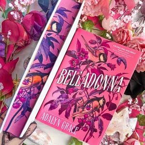 May include: A hardcover book titled "Belladonna" by Adalyn Grace, with a pink cover featuring bird and floral illustrations. The book is open, revealing the spine with matching artwork. Surrounded by pink and purple flowers.
