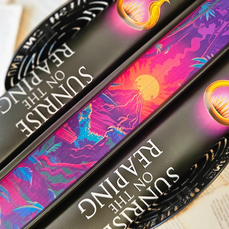 May include: Two hardcover books with the title "Sunrise on the Reaping" on the spine. The top book has a colorful illustration of a sunset with a volcano and palm trees. The bottom book has a gold ring with a sun design.