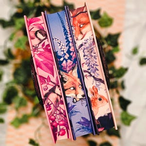 May include: Three decorative books with illustrated covers. The spines are a mix of pink, blue, and purple. The covers feature images of a bird, a fox, and a rabbit, along with floral and botanical elements. The books are arranged in a decorative holder.