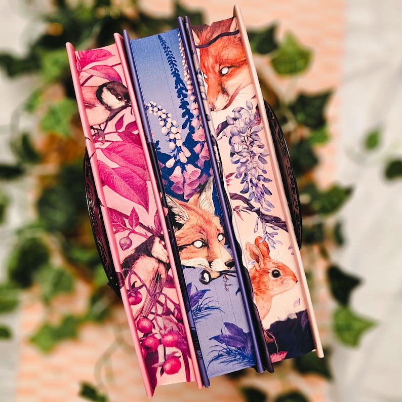 May include: Three decorative books with illustrated covers. The spines are a mix of pink, blue, and purple. The covers feature images of a bird, a fox, and a rabbit, along with floral and botanical elements. The books are arranged in a decorative holder.