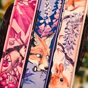 May include: A stack of three decorative books with illustrated covers. The covers feature images of a fox, a bird, and a rabbit, along with floral and botanical designs in shades of pink, purple, and blue. The spines are a deep purple.