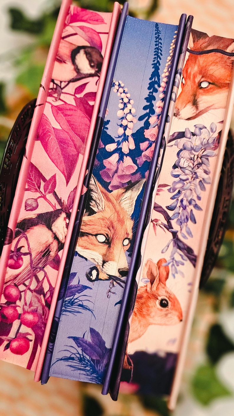 May include: A stack of three decorative books with illustrated covers. The covers feature images of a fox, a bird, and a rabbit, along with floral and botanical designs in shades of pink, purple, and blue. The spines are a deep purple.