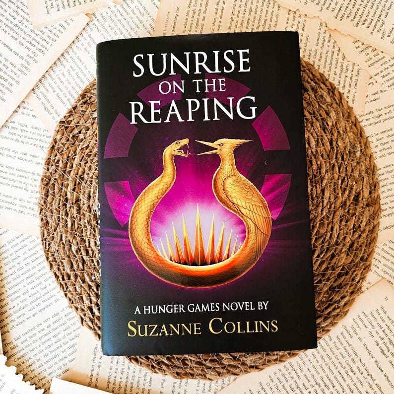 May include: A paperback book titled "Sunrise on the Reaping" by Suzanne Collins. The cover features a purple background with a golden bird and snake design.