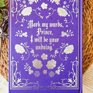 May include: Purple hardcover book with a silver floral design and the text "Mark my words, Prince, I will be your undoing."
