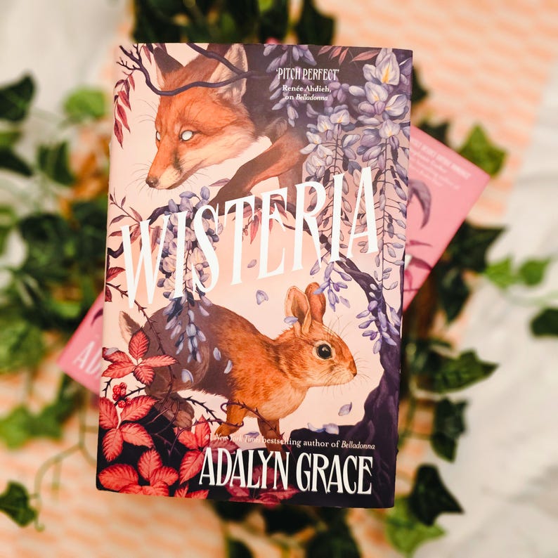 May include: A hardcover book titled "Wisteria" by Adalyn Grace, featuring a fox and a rabbit on the cover. The cover art is a mix of purple, pink, and brown tones, with the title in large white letters. A second book is partially visible.