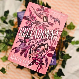 May include: A hardcover book titled "Belladonna" by Adalyn Grace, featuring a pink cover with illustrations of birds, flowers, and foliage. The book has a decorative design with a romantic theme.