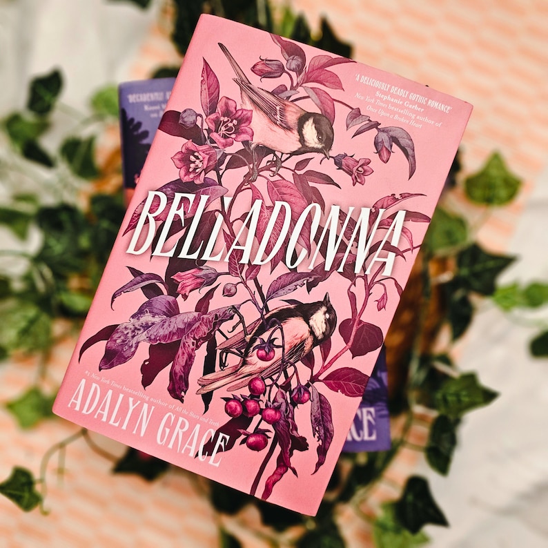 May include: A hardcover book titled "Belladonna" by Adalyn Grace, featuring a pink cover with illustrations of birds, flowers, and foliage. The book has a decorative design with a romantic theme.