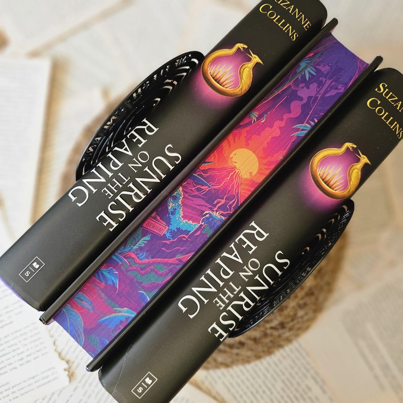 May include: Three black hardcover books stacked on top of each other. The top book is titled "Sunrise on the Reaping" by Suzanne Collins. The middle book is titled "Sunrise on the Reaping" by Suzanne Collins. The bottom book is titled "Sunrise on the Reaping" by Suzanne Collins. The books have colorful covers with a purple, pink, and blue design.