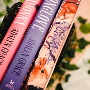 May include: Close-up of three books in a black basket. The books are titled "MADONNA" in pink, "FOXGLOVE" in purple, and a decorative book with a fox and wisteria design. The author's name, Adalyn Grace, is on the spines.