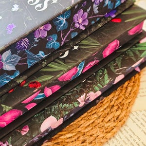 May include: A stack of four hardcover books with floral designs on the covers. The top book has the title "Hunterless" printed on the cover. The books are on a woven surface.