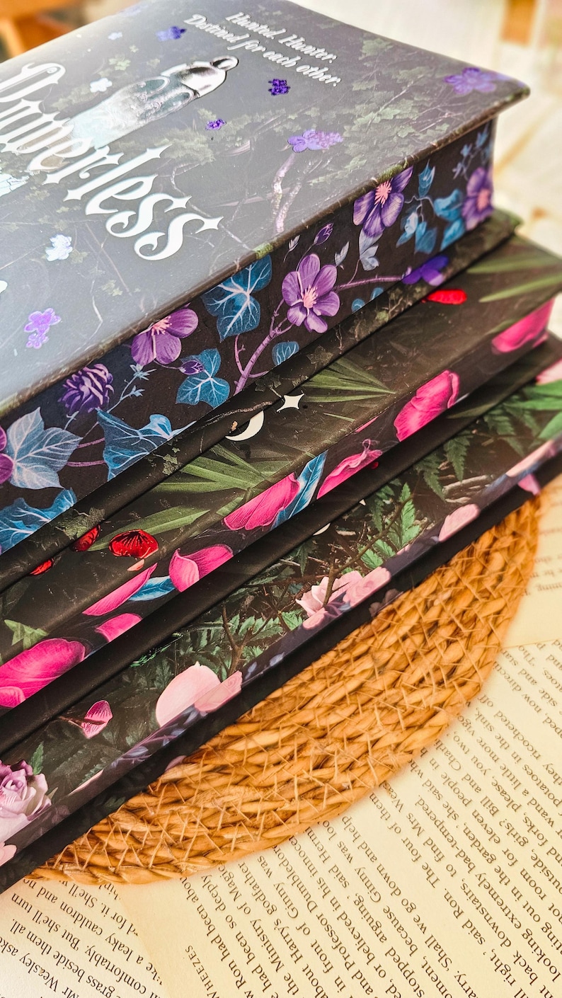 May include: A stack of four hardcover books with floral designs on the covers. The top book has the title "Hunterless" printed on the cover. The books are on a woven surface.