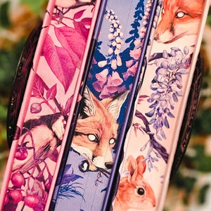 May include: Three decorative books with illustrated covers. The covers feature a fox, a bird, and a squirrel, along with floral and botanical designs in shades of pink, purple, and blue. The spines are a solid purple.