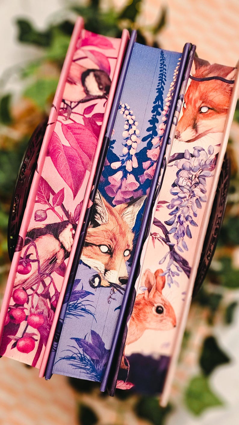 May include: Three decorative books with illustrated covers. The covers feature a fox, a bird, and a squirrel, along with floral and botanical designs in shades of pink, purple, and blue. The spines are a solid purple.