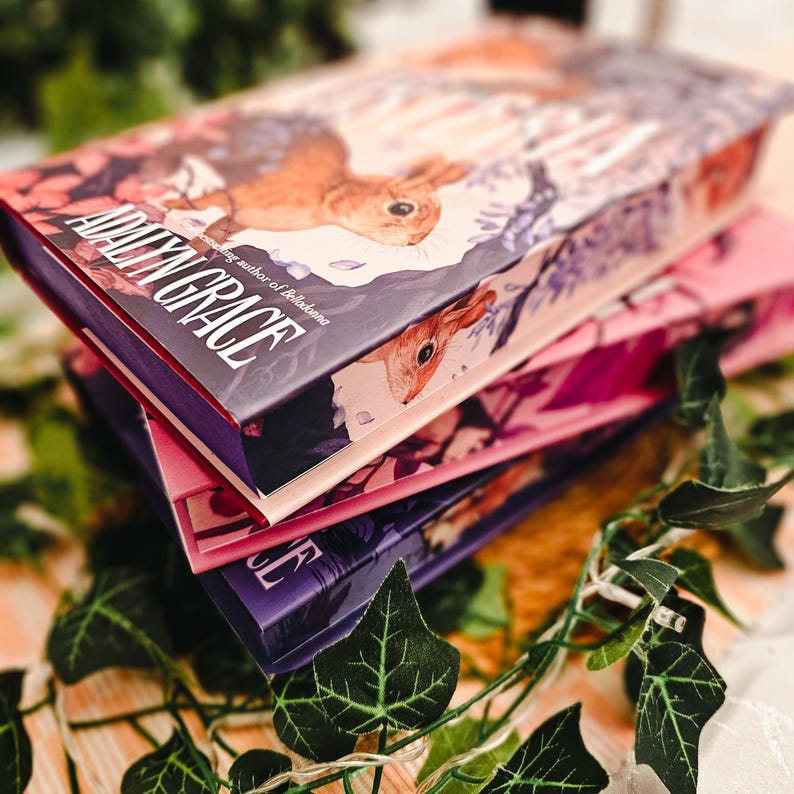 May include: A stack of hardcover books with colorful covers. The top book features the title "Adalyn Grace" with a squirrel illustration and floral accents. The other books are pink and purple. Green ivy vines are draped around the books.