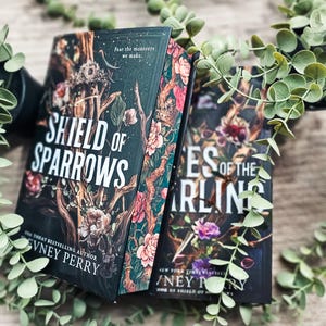 May include: Two hardcover books titled Shield of Sparrows and another partially visible book by Devney Perry. The covers feature dark backgrounds with intricate illustrations of branches, pink and white flowers, and crowns, with the text Fear the monsters we make.