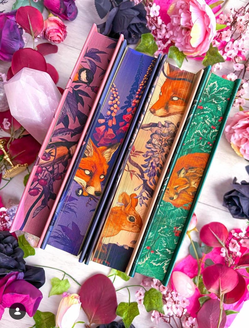 May include: Four decorative books with illustrated covers featuring foxes, flowers, and foliage. The books have pink, purple, tan, and green spines. The image is surrounded by pink and purple flowers.