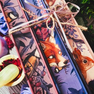 May include: A set of decorative book spines tied with twine and ribbon. The spines feature illustrations of a bird, a fox, and a squirrel, with floral and botanical elements in shades of pink, purple, and blue.