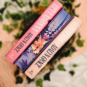 May include: Three hardcover books with the titles "Belladonna" and "Wisteria" are visible. The books are in a black container, with the author's name "Adalyn Grace" printed on the spines. The book covers feature floral and fox illustrations.