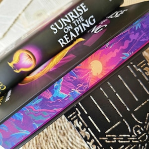 May include: A black hardcover book titled "Sunrise on the Reaping" with a purple and gold design on the cover. The book is stacked on top of another book with a purple, blue, and pink landscape design on the cover.