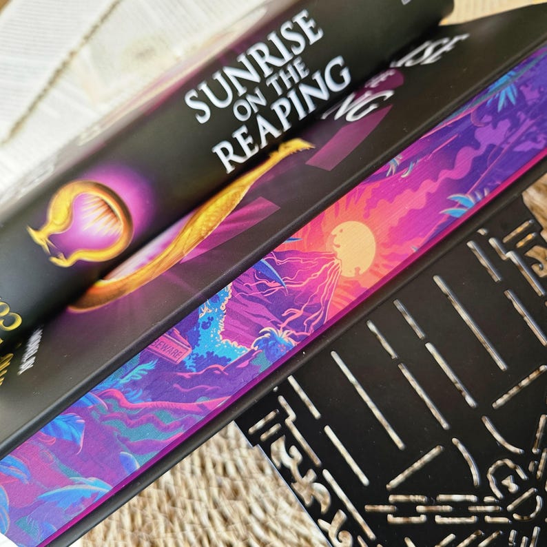 May include: A black hardcover book titled "Sunrise on the Reaping" with a purple and gold design on the cover. The book is stacked on top of another book with a purple, blue, and pink landscape design on the cover.