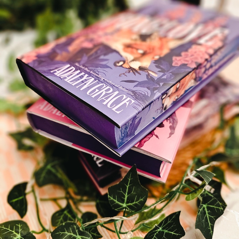 May include: A stack of hardcover books with floral and fantasy illustrations. The top book has a purple cover with the author's name, "Adalyn Grace," in white. The other books have pink and purple covers.