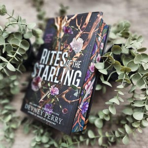 May include: A hardcover book titled Rites of the Starling by Devney Perry. The black cover features a graphic illustration of brown tree branches, white and purple flowers, and green leaves. The title is printed in large white capital letters.