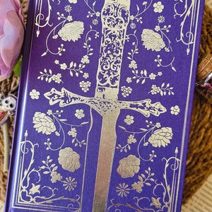 May include: A purple hardcover book with a silver embossed design of a sword surrounded by flowers. The book cover has a floral pattern.