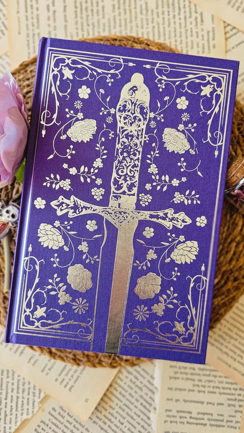 May include: A purple hardcover book with a silver embossed design of a sword surrounded by flowers. The book cover has a floral pattern.