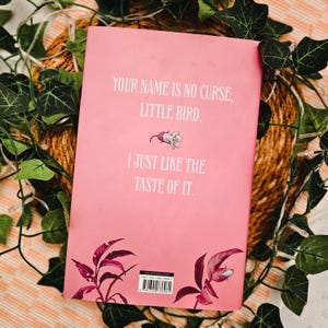 May include: A pink book cover with the text "YOUR NAME IS NO CURSE, LITTLE BIRD. I JUST LIKE THE TASTE OF IT." The cover features a floral illustration and is surrounded by green leaves and a woven background.